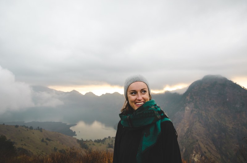 a woman wearing a scarf and a hat smiling atop a cloudy mount rinjani, smiling woman on mount rinjani 4k