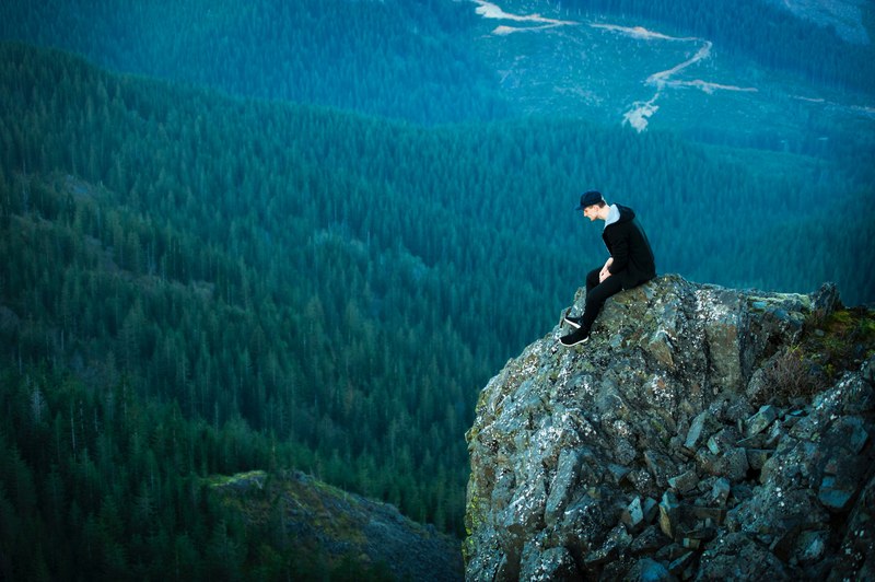 a man in a cap sitting on a ledge overlooking wooded hills, we made it 4k