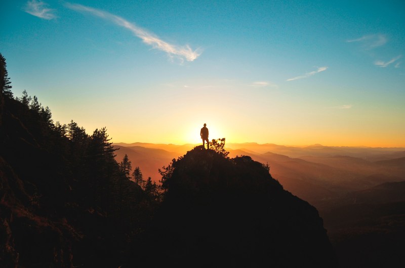 a man standing on a tall rock silhouetted against the setting sun, lost in the wilderness 4k