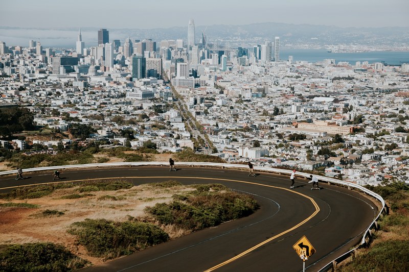 curvy road and kids on skateboard with sprawling cityscape and water behind them, skatin down sf 4k