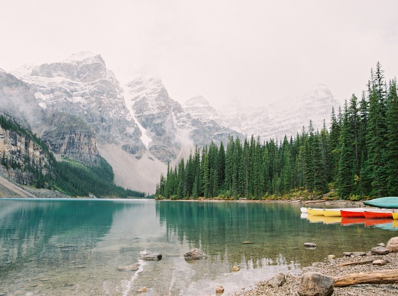 a clear blue lake with colorful canoes lined up on the shore tall evergreen trees line one side while snow capped mountains are visible in the distance, moraine lake 4k