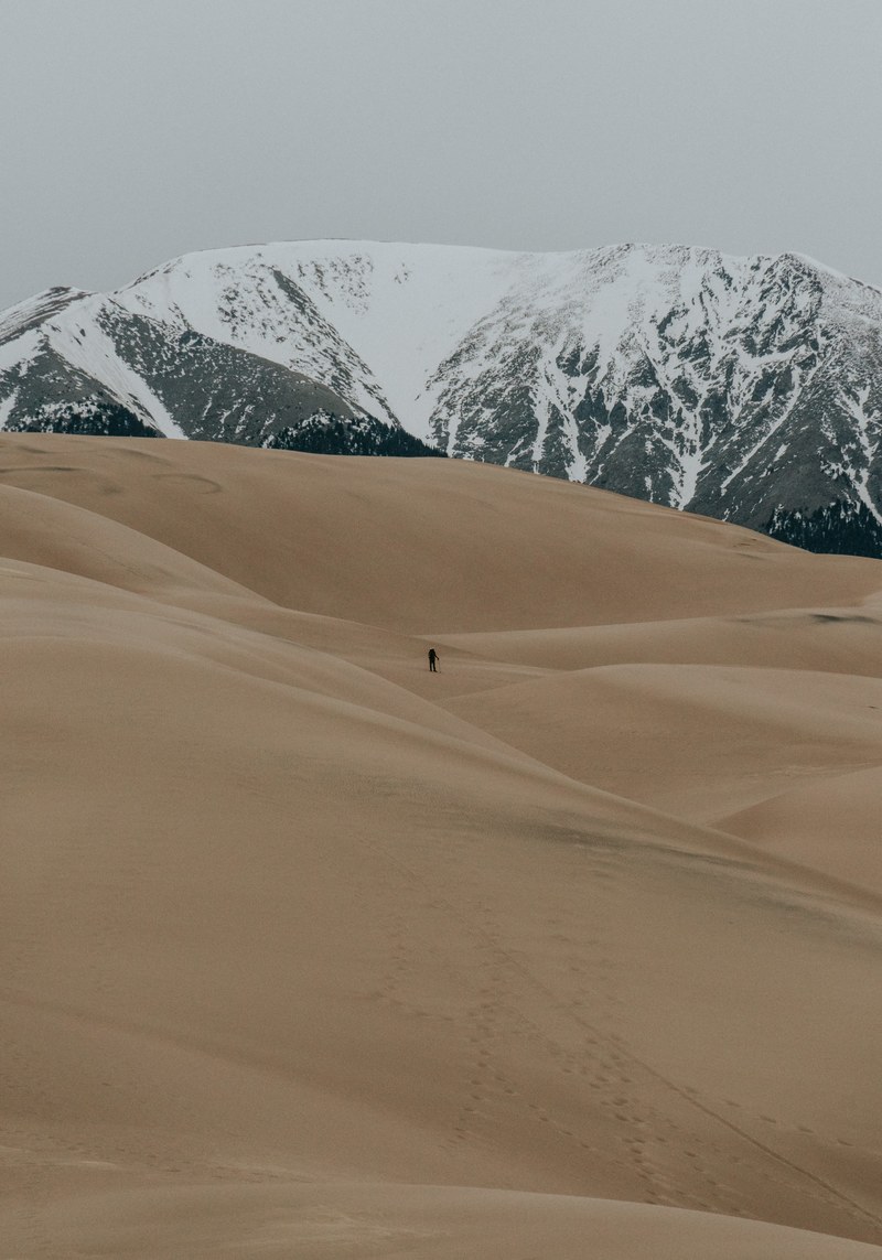 the silhouette of a lone hiker among sand dunes with snowy mountains on the horizon, the great sand dunes 4k