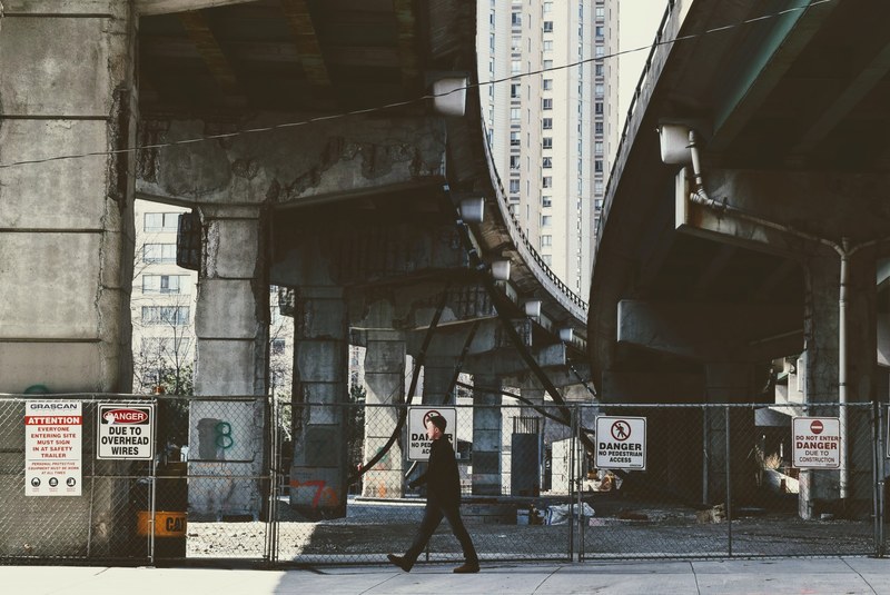 a man walks underneath the overpass along a fenced off area with danger signs, _man walks danger signs 4k