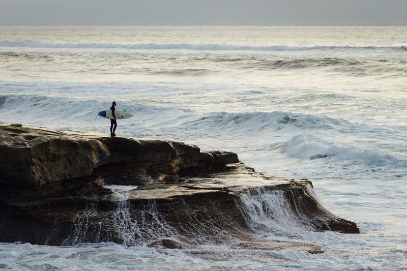 surfer with a surfboard standing on a coastal rock watching rough ocean at ocean beach, sunset surfing 4k