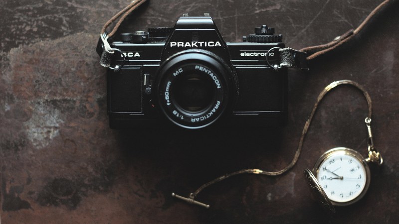 praktica electronic vintage camera with a leather strap beside a gold pocket watch on a scratched brown surface, every day is a new 4k