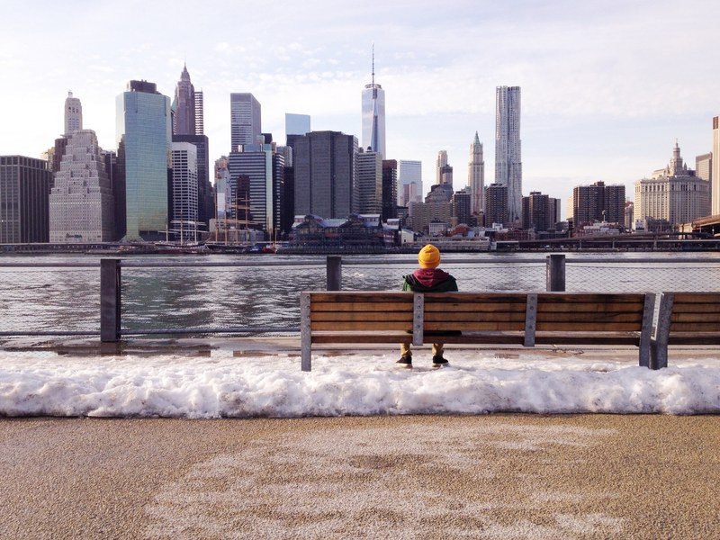 person sits on a bench looking at the water and skyscrapers of new york city, lunch break 4k