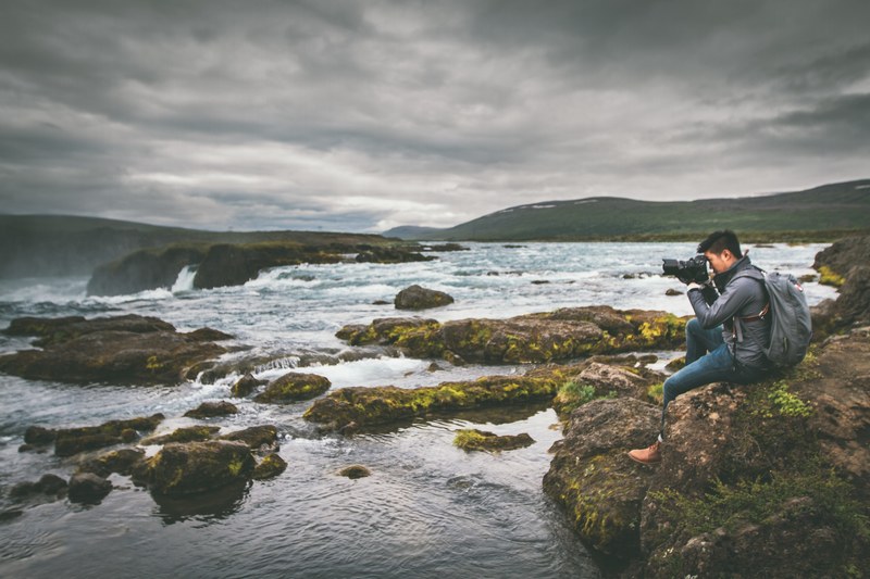 a man sits on a rock near the water to take a picture of the gafoss waterfall, man photographing waterfall 4k
