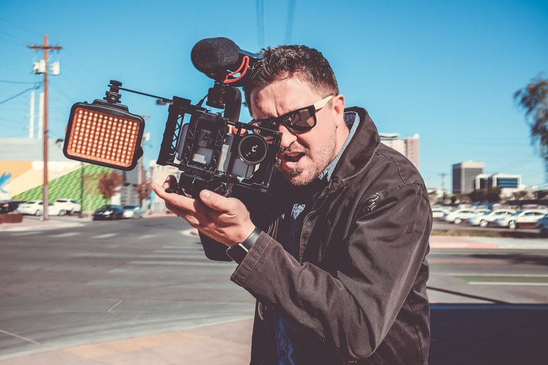 a man in sunglasses holding a professional camera with an excited expression on his face, filming with an iphone in downtown las vegas 4k