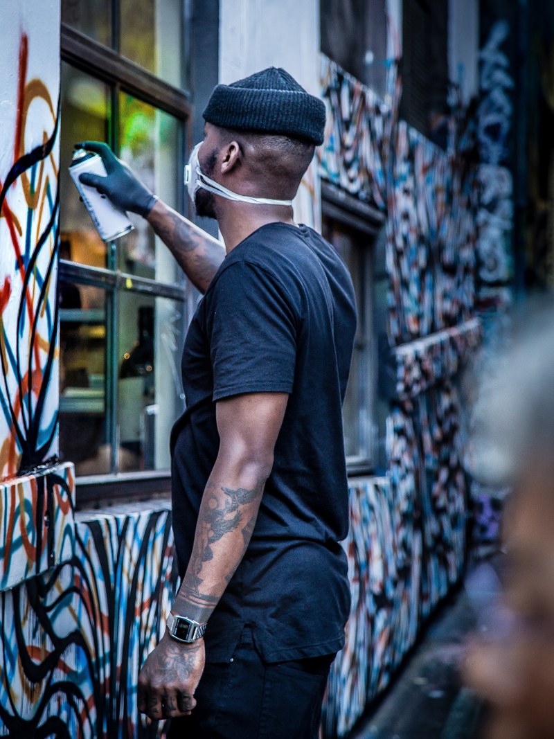 tattooed man wearing a hat and facemask spraypainting graffiti on a building wall on hosier lane, artist spray paints building 4k