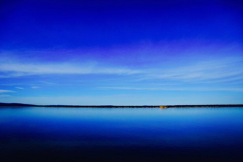 a deep blue lake and clear sky separated by a dark green shoreline on the horizon, blue lake under blue sky 4k