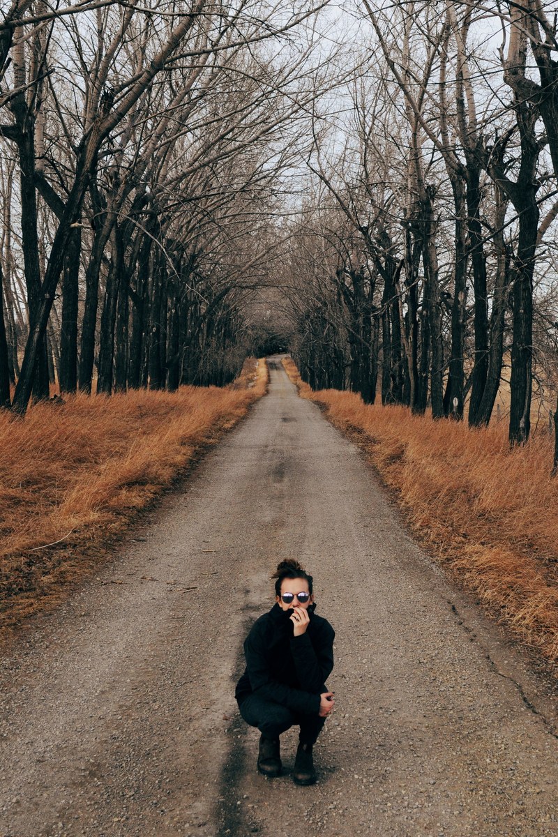 a woman wearing a coat and sunglasses crouched and looking at the camera on a nature path, _woman on path walkway 4k