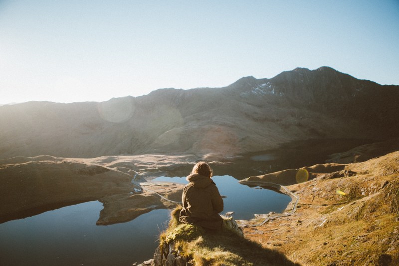 a person sitting on a ledge overlooking a mountain lake near snowdon on a sunny morning, lone wanderer in the mountains 4k