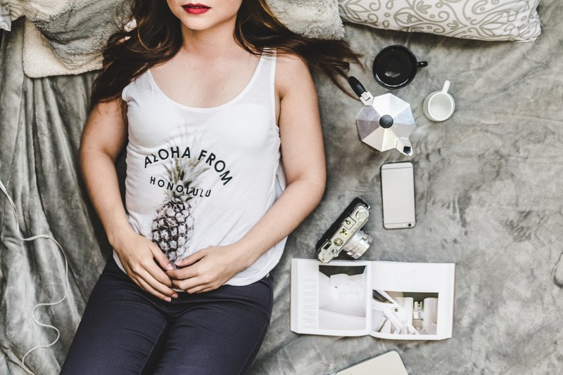 a woman wearing red lipstick and a graphic t shirt laying on a bed with a grey blanket next to a camera iphone cell phone coffee pot coffee cups and book, relax day 4k