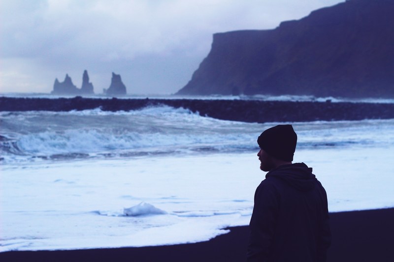 man in a cap looking at the rough ocean on the rocky coastline at vik, man looking at ocean 4k