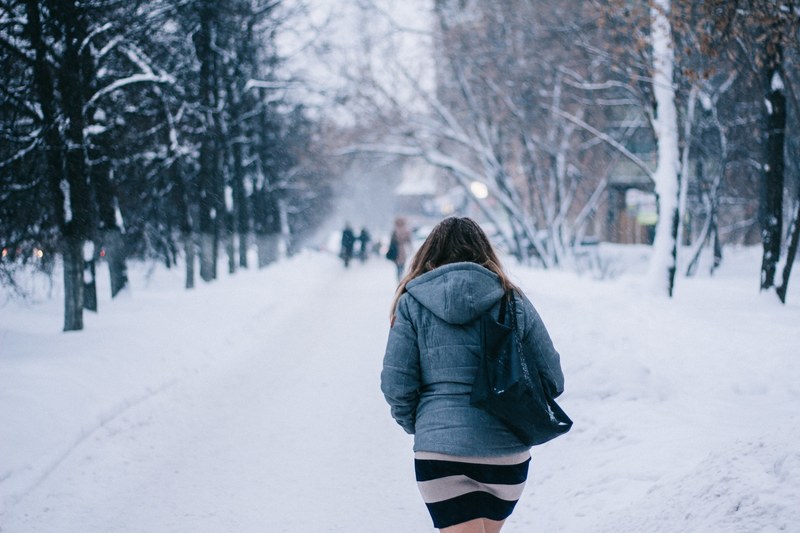 a woman wearing a skirt and jacket walking through the snow on a treed road, woman walking winter road 4k