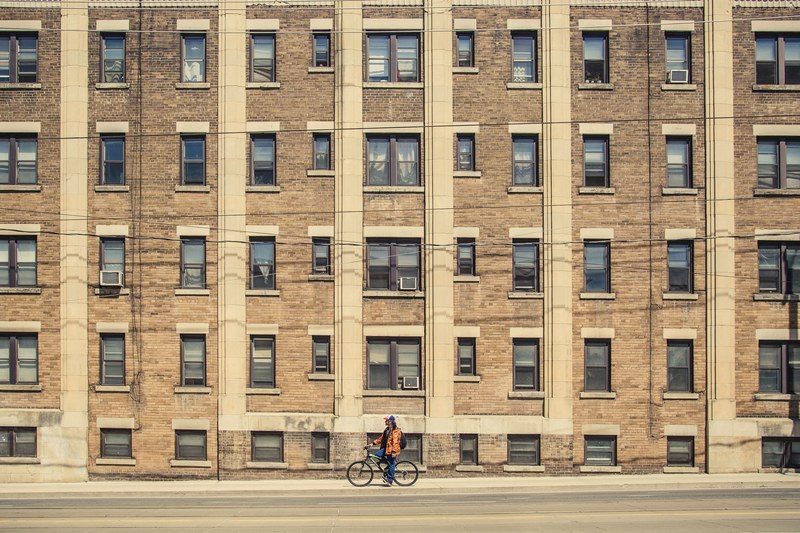 man in orange coat resting on bike in front of brown urban apartment building with windows, man on bike urban apartment 4k