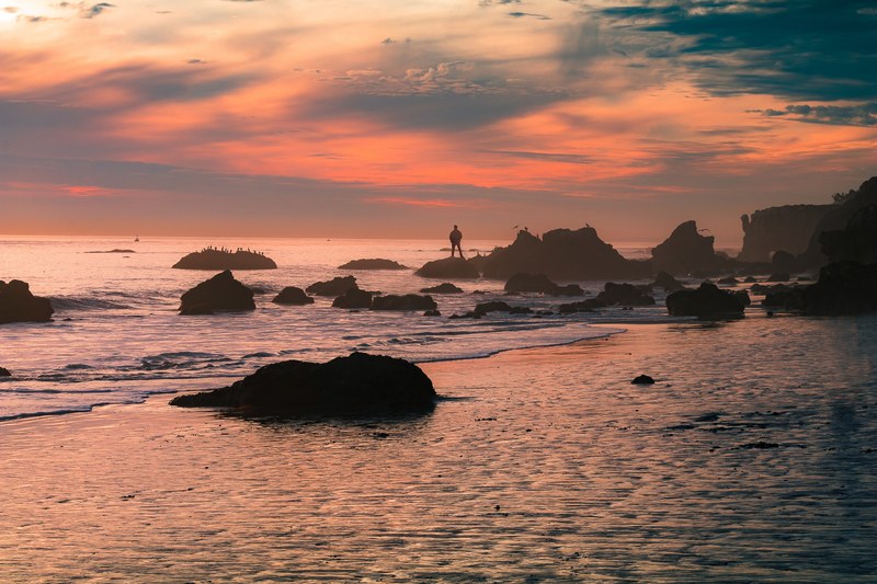 a pastel sunset covers the shores of el matador state beach as a man stands over the rocks, socal tones 4k