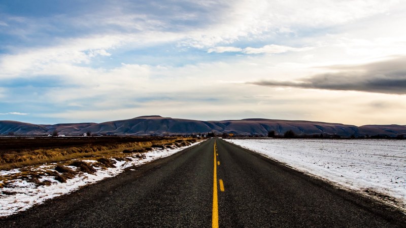 a blacktop road approaches blue mountains and sits between flatland lightly covered in snow, road to mountains 4k