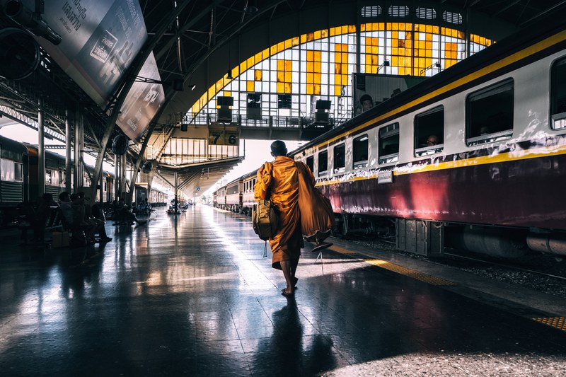a monk in orange robes walking across a railway platform in bangkok, train to catch 4k
