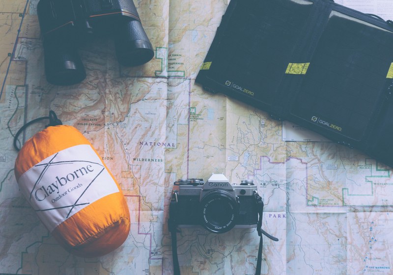 a solar powered portable battery canon camera binoculars and claybourne outdoor goods pack on a map, map flatlay 4k