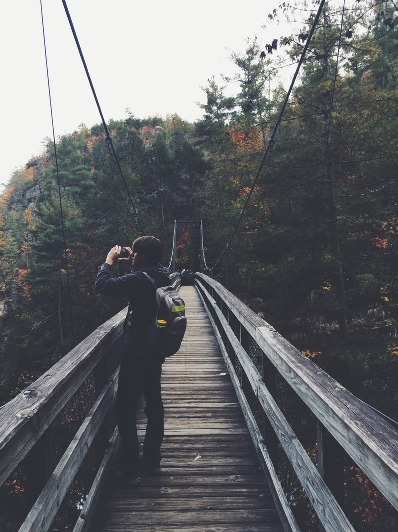 a man in dark casual clothing wearing a gray bag standing on a wooden suspension bridge in an early autumn forest taking a picture of the scenery, hiking instagrams 4k