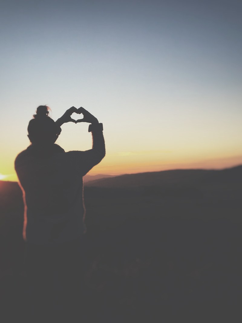 a silhouette of a person making a heart shape with their hands against the twilight sky, heart shape during sunset 4k