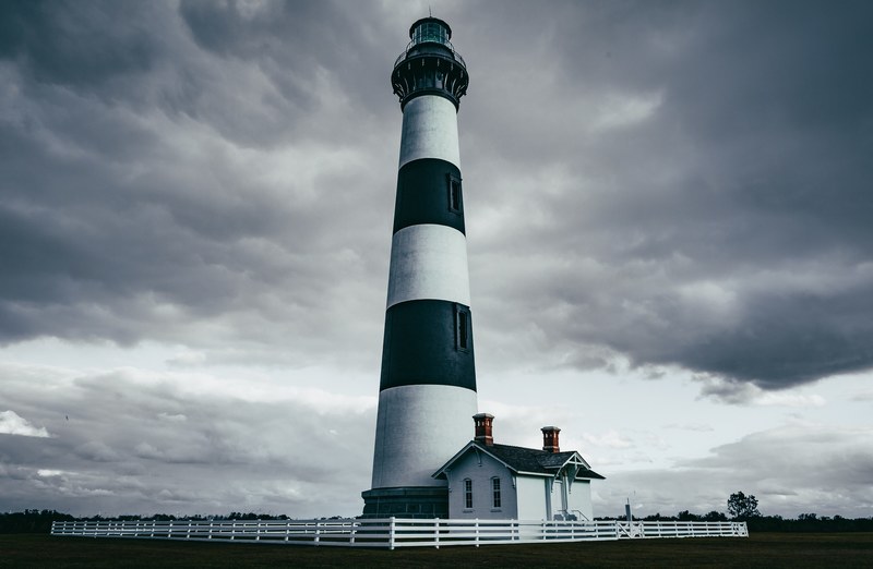 a gray picture of a lighthouse with a small house at the bottom surrounded by a fence, lighthouse on a cloudy day 4k