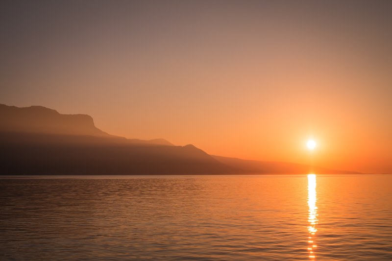 a sunset over a lake next to a silhouette of a steep rock face, sunset by a lake rock face 4k
