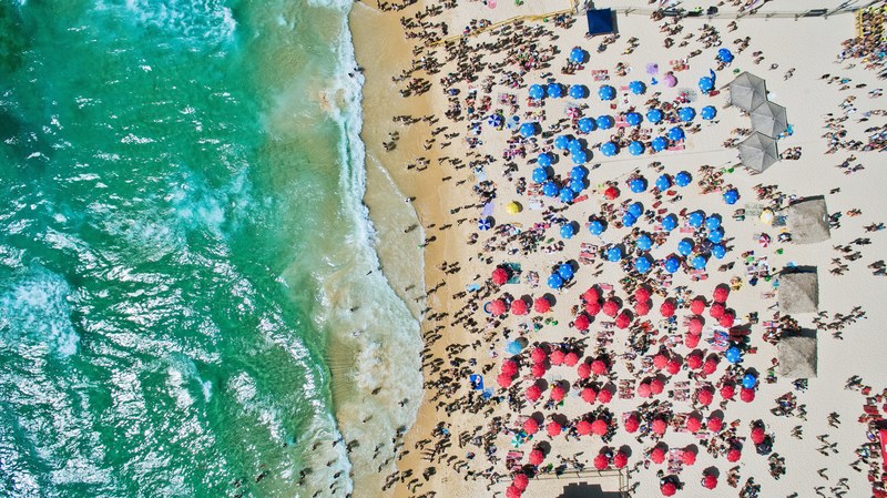 an aerial shot of a crowded beach with vacationers sheltering from the sun under red and blue umbrellas, vertical from above 4k