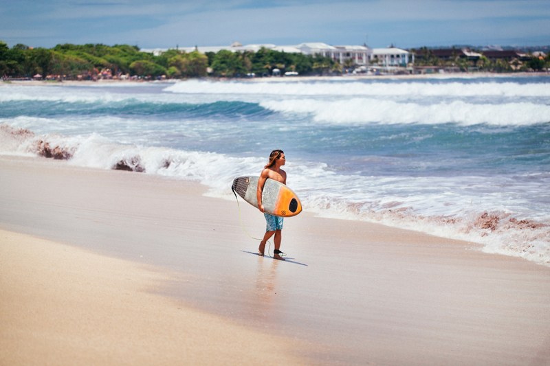 a male surfer carrying a board along a white sandy beach looking at the waves with trees in the distance, surfer at kuta beach bali 4k