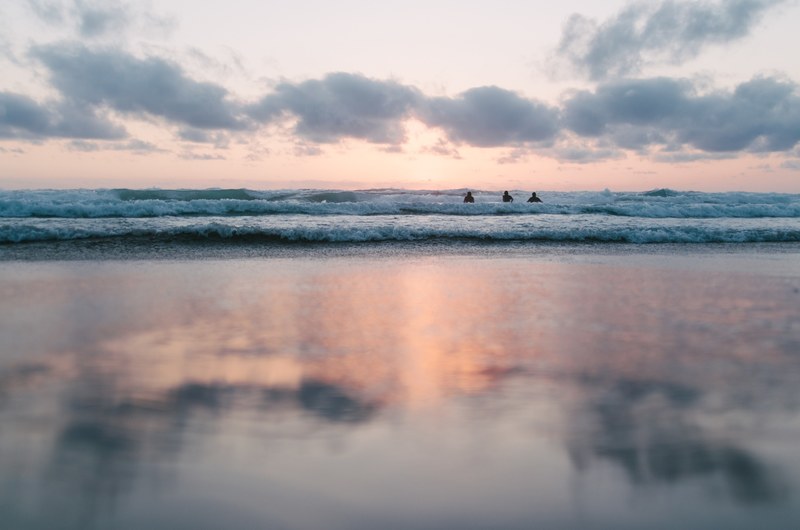 three people in ocean waves with a blurred reflection of a partly cloudy pastel sunset in the foreground, cloud ocean sunset and beach 4k