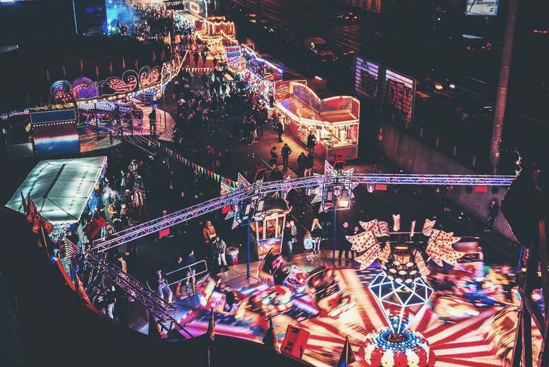 crowd going around a cologne amusement park with rides and stalls lighting up the night, glowing amusement park lights 4k