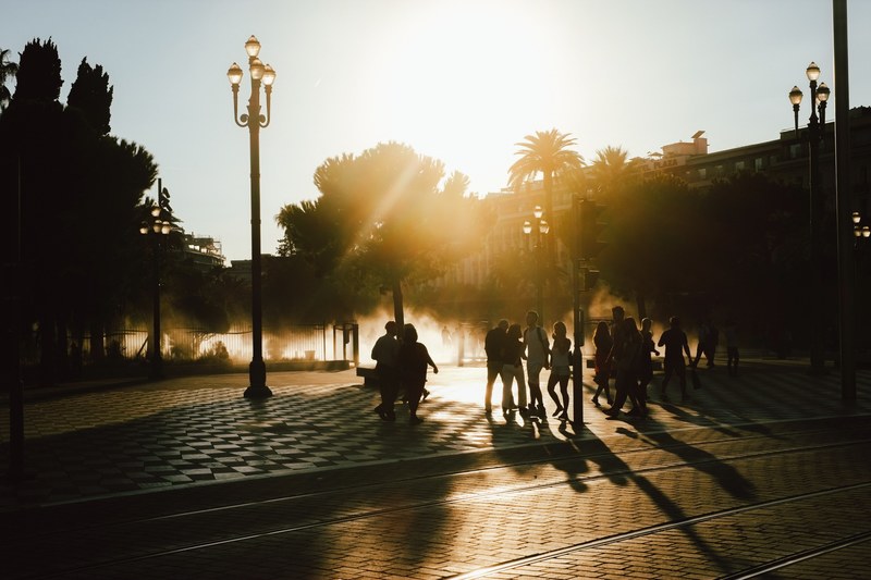 silhouettes of pedestrians on the streets of nice as the sun sets behind palm trees in the distance, pedestrians at sunset 4k