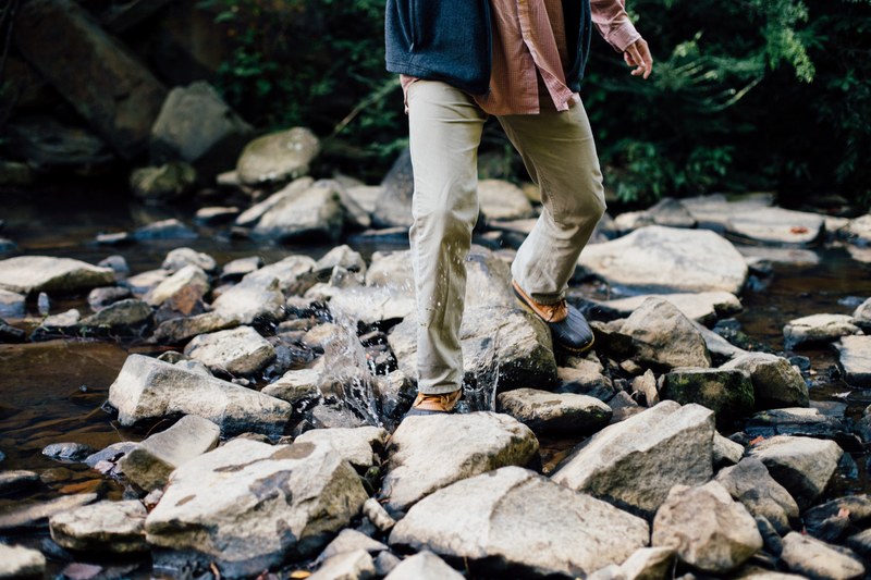 a man splashing across a stream in duck boots and a vest, splashing water between rocks 4k
