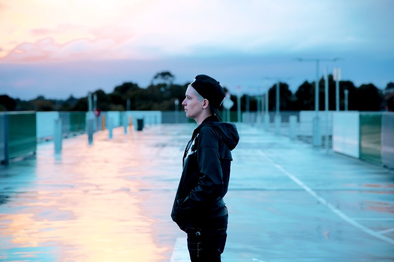 a person in a backwards baseball cap and black clothing stands on sunny pavement, person on sunny pavement 4k