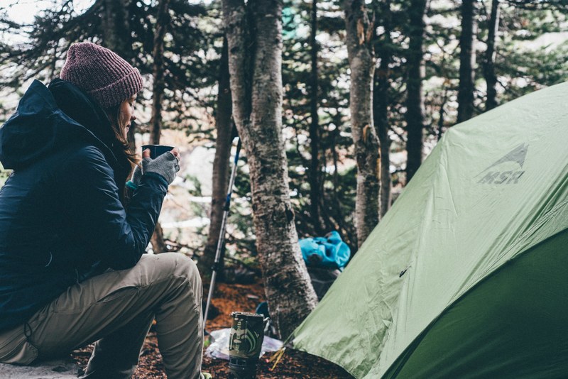a camping woman enjoying her tea near a tent under the trees on a cold day, female camper enjoying morning tea 4k