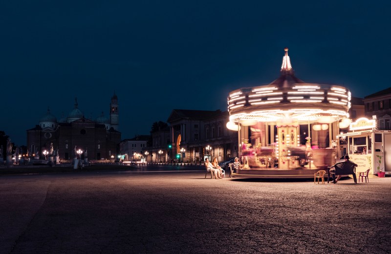 adults watching a bright spinning carousel in the streets of prato della valle at night, spinning carousel in the night 4k