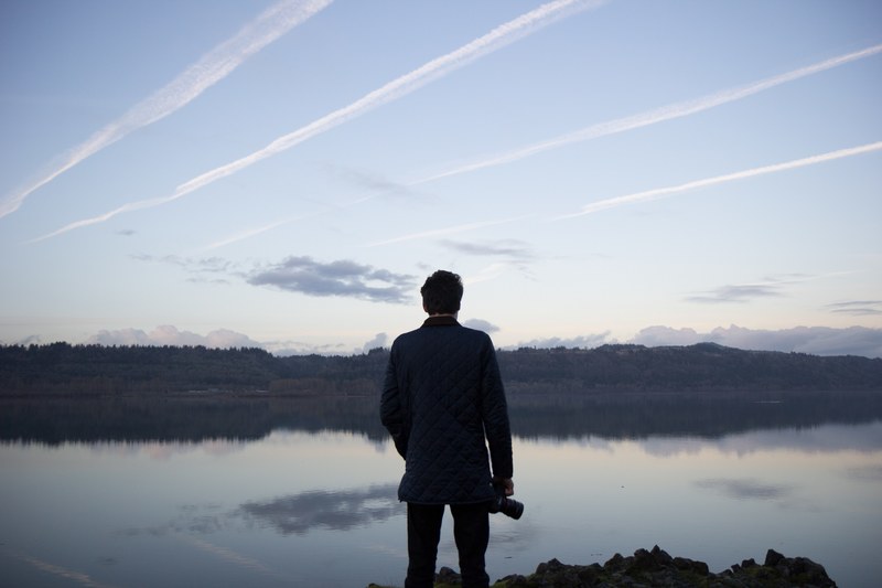 a photographer holding their camera looking out at the calm water and forest, photographer and lake 4k