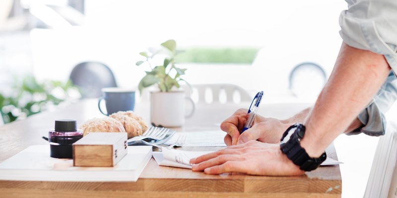 a standing man writing on sheets of paper on a desk with a cup and pastries, man writing while standing 4k