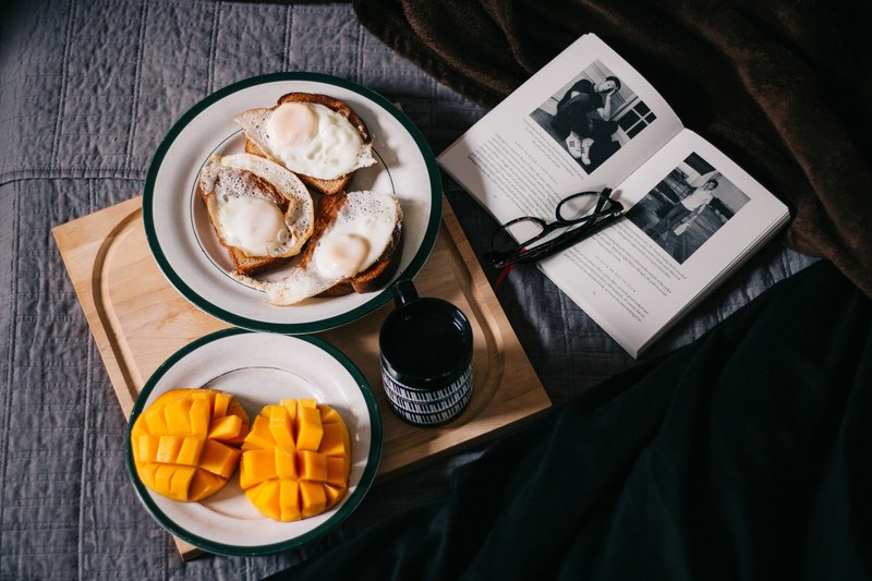 a breakfast spread including mango fried eggs and coffee on a wooden serving board next to a book and glasses on a bed, breakfast in bed 4k