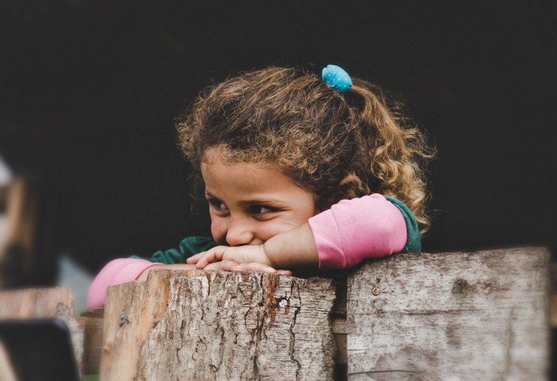 a curly haired young girl places her head on her crossed arms on a wooden wall, smile of life 4k