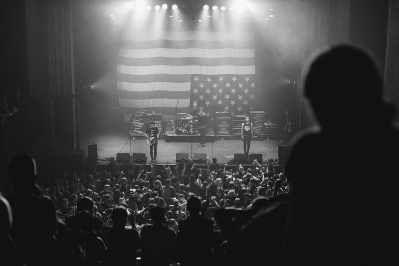 a black and white shot of a rock band performing in front of a large upside down american flag, anti flag montreal january 2017_ 4k