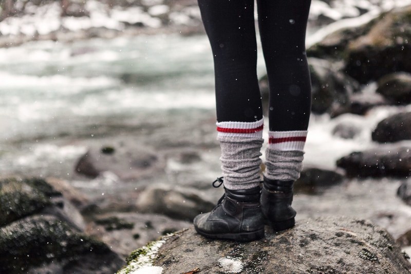 a person with leggings black boots and bunched socks stands on a rock during a light snow only the persons legs are in frame, boots on a rock 4k