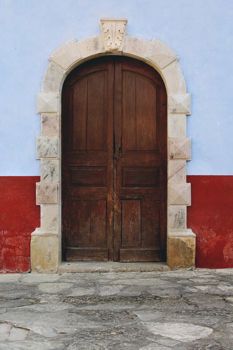 large domed wooden door with white stones on the border and a red and blue wall flanking it on a stone street, red and blue wall with wood door 4k