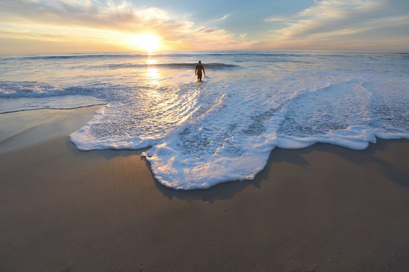 man entering the foamy ocean from the sand beach at sunset, surfer at sundown 4k