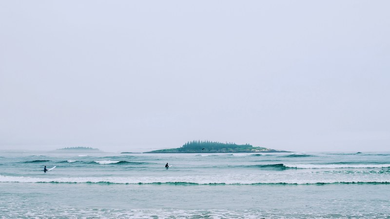 two surfers in the sea at popham beach with a tree covered beach or island in the background, surfing on an overcast day 4k