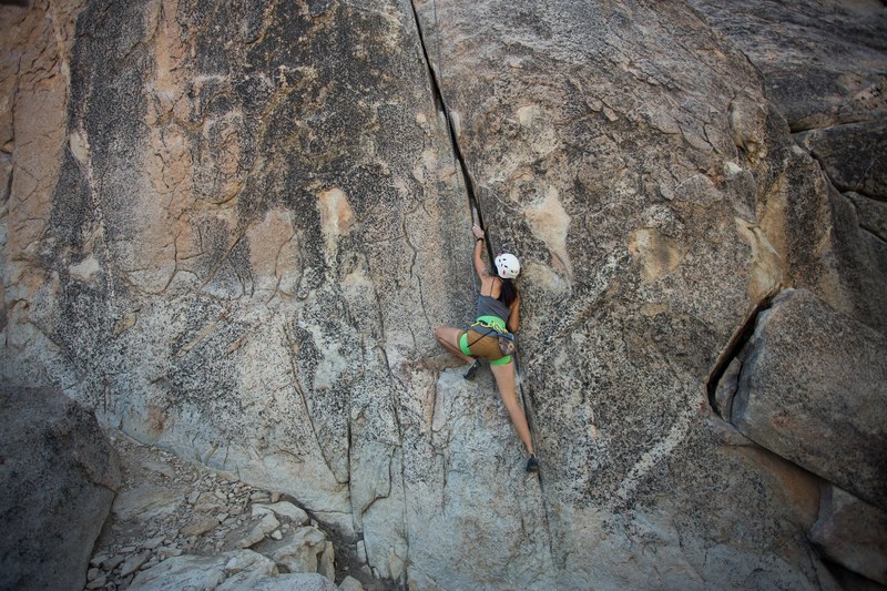 a woman wearing a white helmet and green shorts rock climbing the side of a mountain in big bear lake, casey climbing 4k