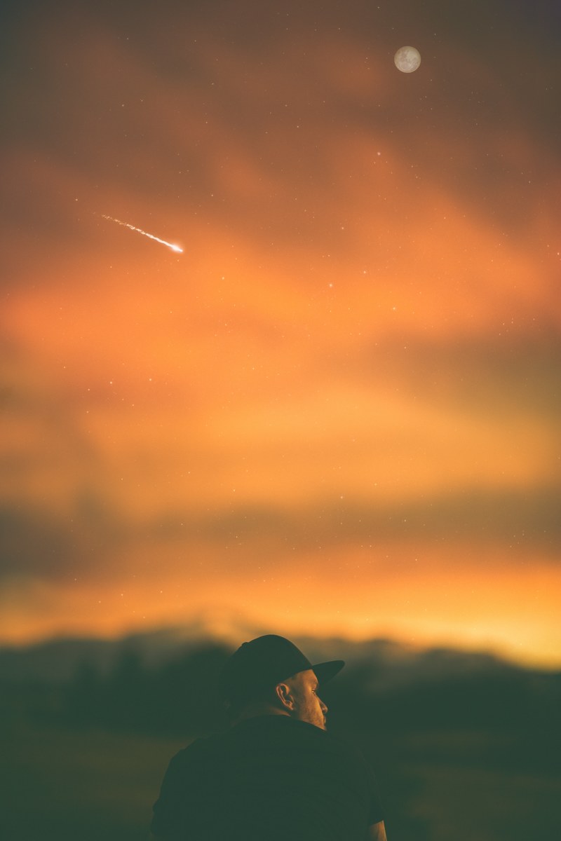 man sits beneath an orange sky where a meteor streaks and a full moon rises, man moon meteor at dusk 4k