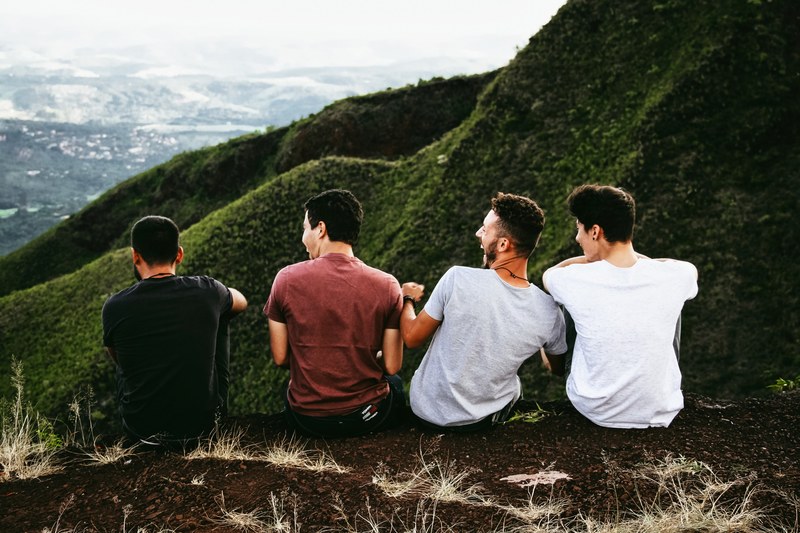 a group of four laughing men sits on an edge overlooking a green valley, men laughing by green valley 4k