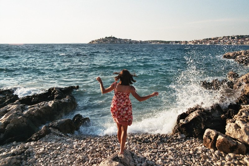 a woman in a dress standing on a rock on the coast with waves crashing against the nearby rocks, sea breeze and splashing waves 4k